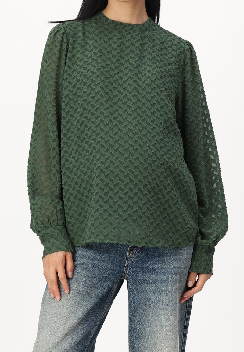 Blouse verte texturée avec un motif en chevron, manches longues et col rond, associée à un jean en denim bleu clair.