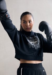 Sweatshirt noir court avec logo blanc, associé à un short noir avec des rayures blanches sur les côtés. Gants de boxe noirs avec des accents en camouflage.