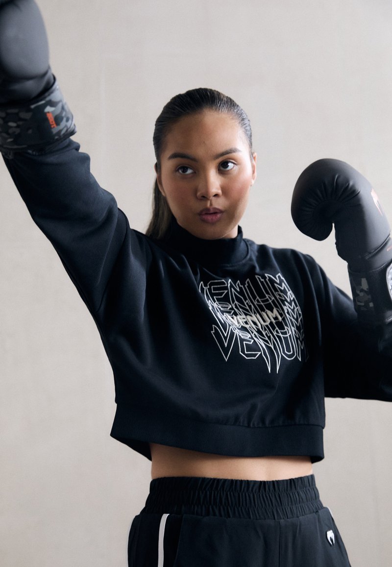 Svart kort sweatshirt med vit logotyp, kombinerad med svarta shorts med vita sidostrecker. Svarta boxningshandskar med kamouflage-accenter.