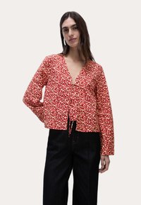 Rød bluse med blomstermønster, v-hals, lange ærmer og en cropped design. Har en snøre lukning i taljen og er lavet af letvægtstof.