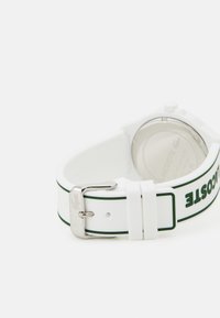 White silicone watch strap with green accent stripes, silver buckle, and embossed logo. Round silver watch case with a clear back.