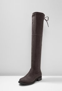 Bota alta de ante marrón oscuro por encima de la rodilla con tacón cuadrado y un lazo decorativo en la parte trasera superior, mostrada sobre una superficie blanca y con un fondo gris.