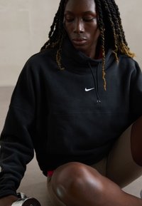 Ung kvinde med fletninger iført en sort Nike hættetrøje, siddende og holder en sølv håndvægt på en neutral baggrund.