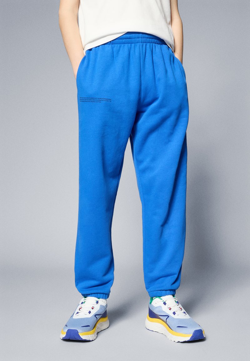 Pantalones de chándal azules con cinturilla elástica y bolsillos laterales. Detalle de texto en la parte trasera. Combinados con zapatillas multicolores que presentan un diseño de malla y goma.