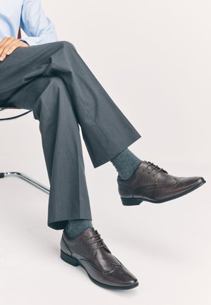 Braune Lederschuhe mit perforierten Brogue-Details, kombiniert mit grauen, strukturierten Socken und maßgeschneiderten grauen Hosen, sitzend auf einem Stuhl aus Chrom.