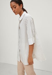 Camicia a righe bianca e marrone chiaro, con maniche lunghe arrotolate, colletto e vestibilità rilassata. Tessuto dalla texture liscia.