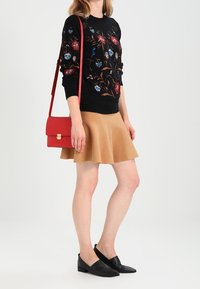 Maglione nero con ricami floreali, gonna svasata beige, borsa a tracolla rossa e scarpe slip-on nere. Mix di textures e colori.