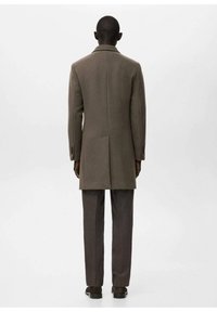 Abrigo de lana marrón con corte recto, cuello con muesca y aberturas laterales. Combinado con pantalones marrón oscuro y zapatos negros, visto desde atrás.