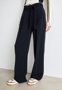 Pantaloni palazzo blu navy con vita legata, parte frontale plissettata e texture liscia, abbinati a sneakers bianche e beige.