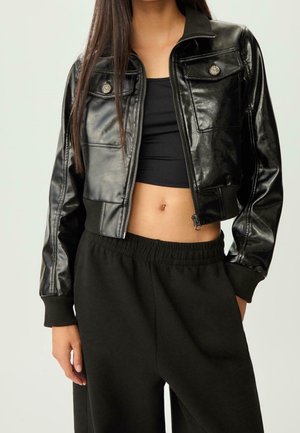 Femme portant une veste courte en cuir noir sur un crop top noir, associée à un pantalon noir à taille élastique, debout avec une main dans la poche.