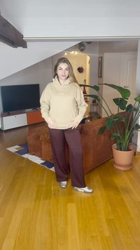 Sweatshirt bege com capuz e bolso frontal, combinado com calças de perna larga em bordô escuro. Ténis desportivos prateados. Ambiente interior com plantas.