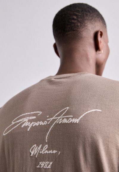 Tricou tricotat maro, având un mare logo alb scris de mână pe spate, cu detalii de text "Giovanni Arnumo, Milano, 1982".