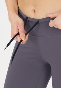 Grå sportleggings med en slät yta, med en svart dragsko i midjan. Leggingsen har en figurnära design och synliga sömdetaljer.