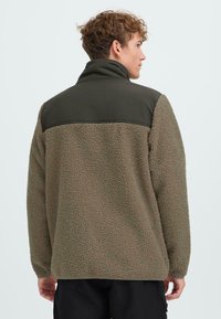 Giacca a pullover in pile marrone con una parte superiore liscia di colore verde scuro. Presenta un colletto alto e un tessuto texturizzato, che offre calore e comfort.