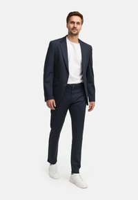 Completo blu con giacca monopetto e pantaloni su misura, abbinato a una t-shirt bianca e sneakers bianche. Tessuto liscio, vestibilità moderna.