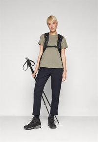 Olivinės spalvos marškinėliai, juodas reguliuojamas kuprinė, juodos kelnės ir žygių batai. Laiko žygių lazdas. Paprastas dizainas; praktiška lauko apranga.