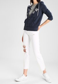 Marineblauwe zip-up hoodie met witte trekkoorden, gecombineerd met versleten witte jeans met rafelige zoom, en witte sneakers.