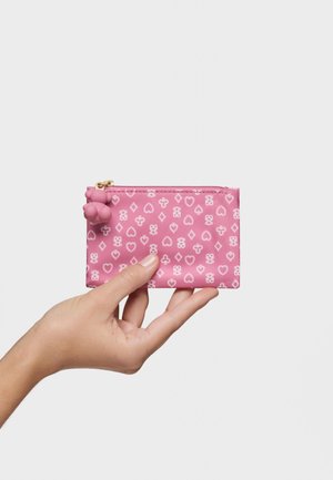 Una mano sostiene una pequeña bolsa con cremallera rosa con patrones de corazones, diamantes y tréboles blancos, y un tirador de cremallera en forma de oso.