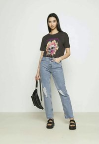 Jovem mulher vestindo uma t-shirt gráfica preta, jeans claros rasgados, sandálias pretas, segurando uma bolsa preta, de pé contra um fundo claro simples.