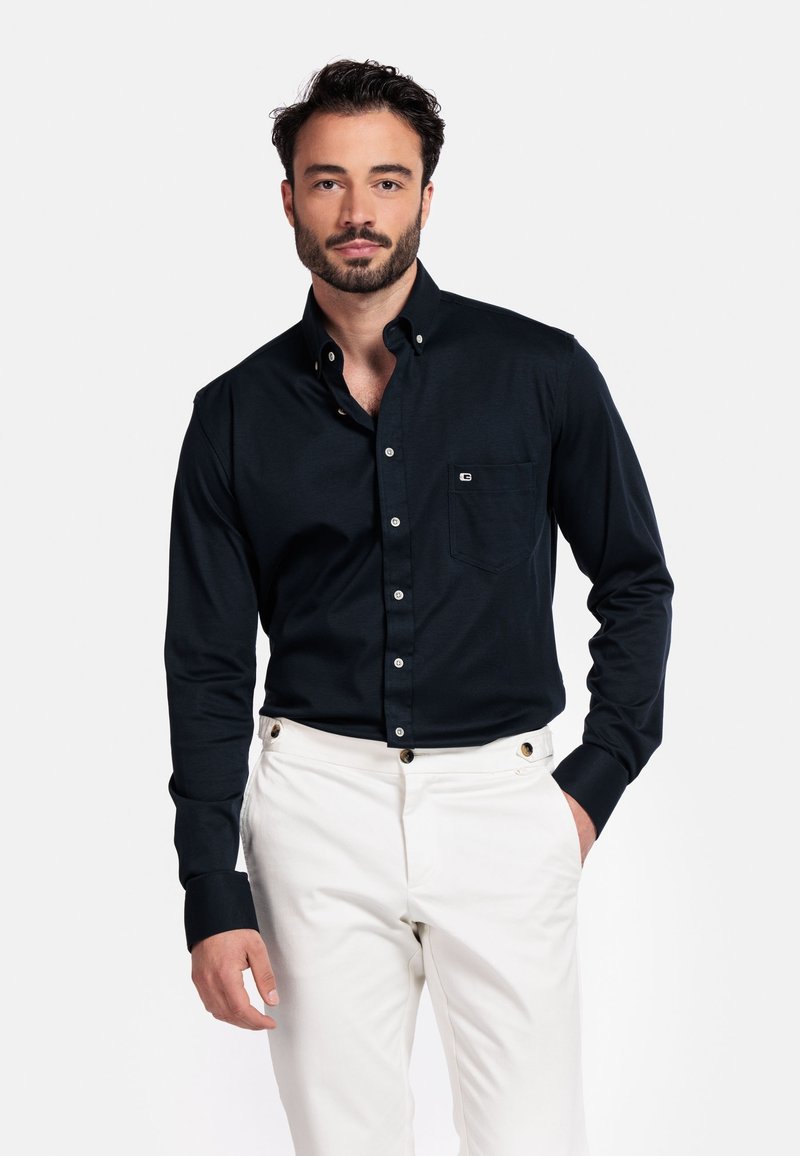 Man met kort donker haar en baard, gekleed in een zwart overhemd met knopen en witte broek, staand met één hand in zijn zak tegen een witte achtergrond.