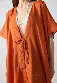 Cubierta ligera naranja hecha de tejido texturizado, con escote en V, mangas cortas y cintura atada para un ajuste ajustable.