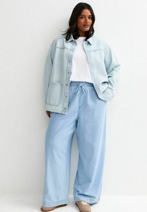 Mujer con chaqueta de mezclilla azul claro y pantalones de pierna ancha, camiseta blanca, zapatos beige, de pie contra un fondo blanco liso.