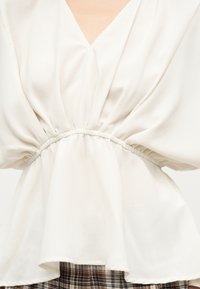 Blouse blanche avec taille froncée et col en V, portée sur un pantalon à carreaux marron et blanc.