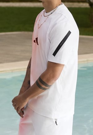 Henkilö, jolla on valkoinen Adidas-t-paita, jossa on musta vinoraita hihassa, ja kaksi mustaa kyynärvarren tatuointia, seisoo uima-altaan vieressä.