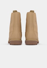 Une paire de bottes en cuir beige vues de l'arrière, montrant des détails cousus et un logo en relief sur le côté.