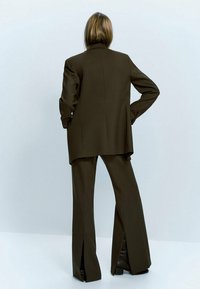 Blazer marron ajusté avec épaulettes et pantalons évasés assortis avec fentes avant. Texture lisse, coupe structurée, détails minimalistes.