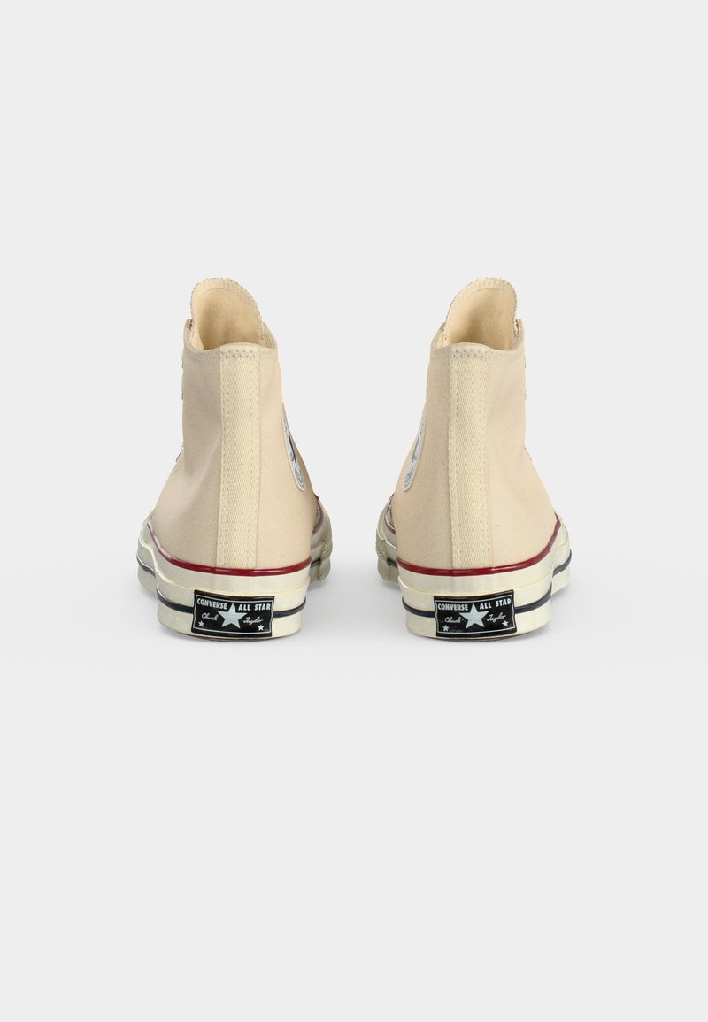 Ein Paar beige Converse All Star High-Top-Sneaker, von hinten gezeigt, mit sichtbaren schwarzen Fersenetiketten und rotem Streifen auf weißen Sohlen.