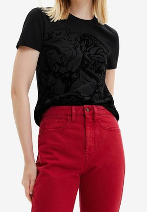 Nainen, joka käyttää mustaa graafista t-paitaa ja punaisia korkeavyötäröisiä farkkuja, seisoo yhdellä kädellä rentona sivulla.