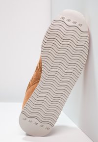 Vaaleanruskea mokkanahkainen tennari, jossa valkoinen kumipohja, jossa aaltoileva uritettu kuvio ja pyöristetty varvasmuotoilu. Koristeina ommellut yksityiskohdat.