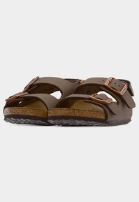 Καφέ δερμάτινα σανδάλια με δύο ρυθμιζόμενες αγκράφες σε χαλκοχρώμιο και πάτο από φελλό, απεικονιζόμενα από χαμηλή μπροστινή γωνία σε λευκό φόντο.