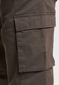 Pantalones cargo marrón oscuro con una tela texturizada, que presentan un gran bolsillo lateral y detalles de costura visibles. Diseñados para la practicidad.