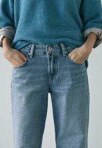 Person wearing light blue denim jeans with hands in pockets and a blue long-sleeve sweater with rolled-up cuffs.