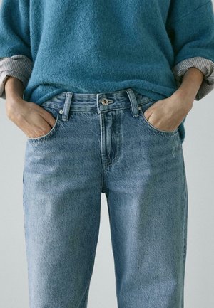 Persona che indossa jeans di denim azzurro chiaro con le mani nelle tasche e un maglione a maniche lunghe blu con polsini arrotolati.
