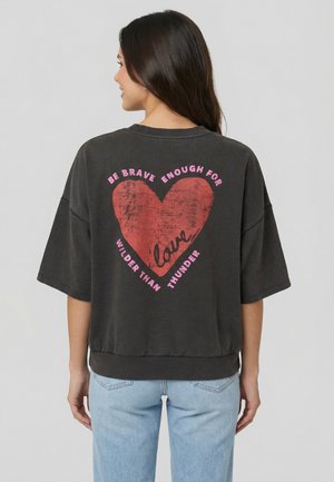 Femme aux longs cheveux foncés portant une chemise noire oversize avec un grand cœur rouge et un texte rose dans le dos, associée à un jean bleu clair.