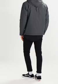 Grå udendørsjakke med hætte, elastiske manchetter og skulderlap, parret med sorte bukser og sorte sneakers med hvide såler og sokker.