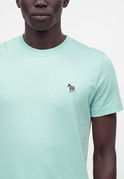 Άνδρας που φοράει ένα ανοιχτοπράσινο t-shirt με στρογγυλή λαιμόκοψη και μια μικρή πολύχρωμη κεντημένη ζέβρα στο αριστερό στήθος.