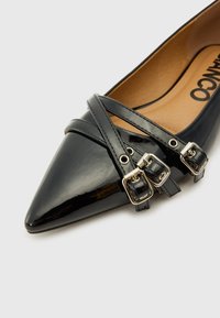 Una scarpa flat in vernice nera con punta affusolata, caratterizzata da cinturini incrociati e fibbie metalliche per decorazione.
