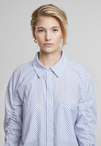 Camicia oversize a righe blu e bianche con collo, chiusura con bottoni e due tasche frontali. Tessuto morbido con maniche testurizzate.