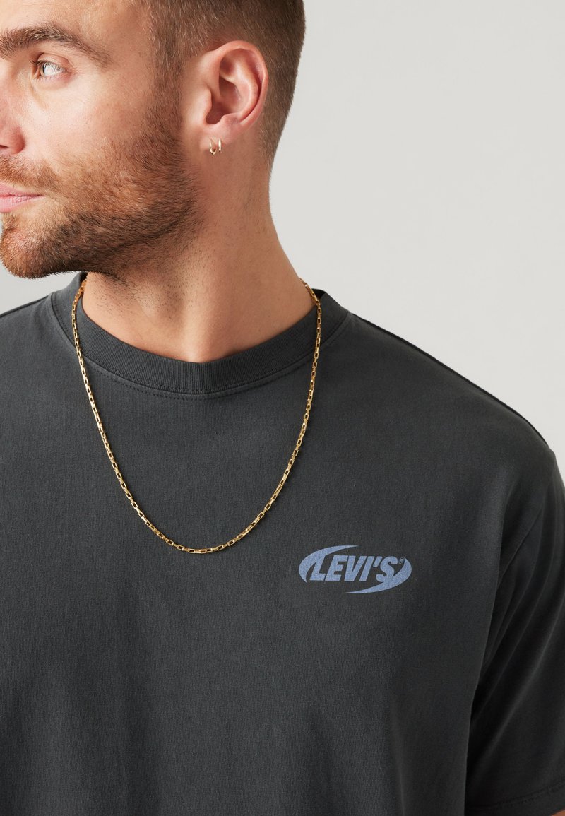 Bärtiger Mann mit kleinen goldenen Creolen, einer goldenen Halskette und einem schwarzen Levi's T-Shirt, der nach links schaut.