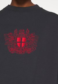 Sudadera gris oscuro con un escudo bordado en rojo que muestra un escudo con una cruz y aves estilizadas a cada lado.