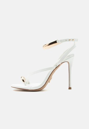 Λευκή σανδάλι με ψηλό τακούνι και λεπτά λουράκια, διακοσμητικό χρυσό δαχτυλίδι στα δάχτυλα και χρυσή αγκράφα στο λουράκι του αστραγάλου, απεικονίζεται σε πλευρική όψη.