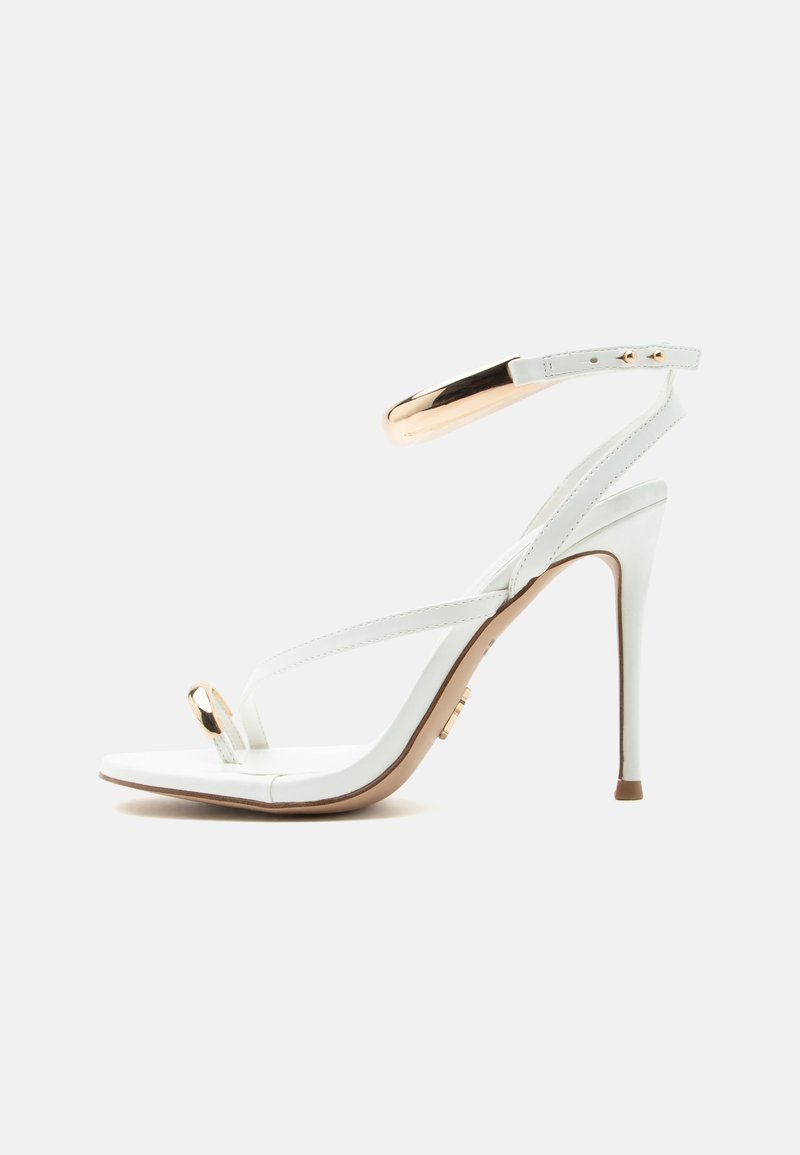Λευκή σανδάλι με ψηλό τακούνι και λεπτά λουράκια, διακοσμητικό χρυσό δαχτυλίδι στα δάχτυλα και χρυσή αγκράφα στο λουράκι του αστραγάλου, απεικονίζεται σε πλευρική όψη.