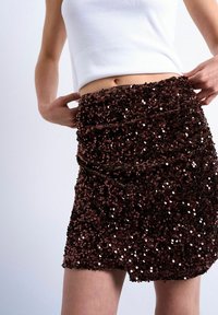 Femme ajustant une mini-jupe taille haute en sequins brun foncé, portant un top blanc sans manches, sur un fond clair uni.