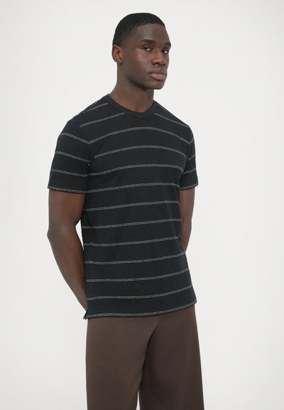 Schwarzes T-Shirt mit weißen horizontalen Streifen, kurzen Ärmeln und Rundhalsausschnitt. Getragen mit braunen Hosen in entspannter Passform.