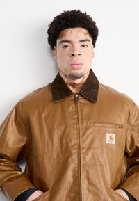 Giacca rivestita in marrone con collo marrone scuro, tasca con zip e patch del logo. Il tessuto è liscio con una leggera texture increspata.
