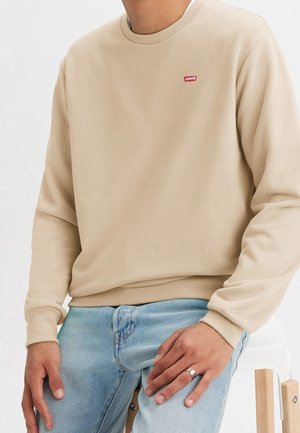 Osoba ubrana w beżową bluzę z okrągłym dekoltem z czerwonym logo Levi's i jasnoniebieskie dżinsy, siedząca na białym stołku.